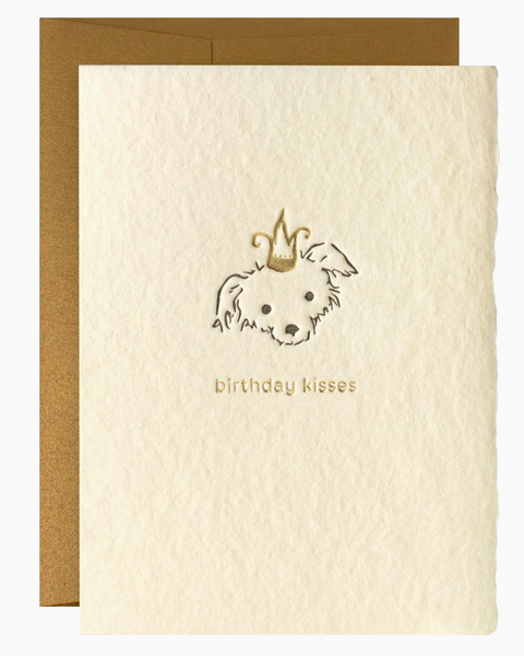 A cream-colored textured greeting card with a gold foil illustration of a dog wearing a crown and the words 'birthday kisses' below, shown with a gold envelope behind it on a white background.